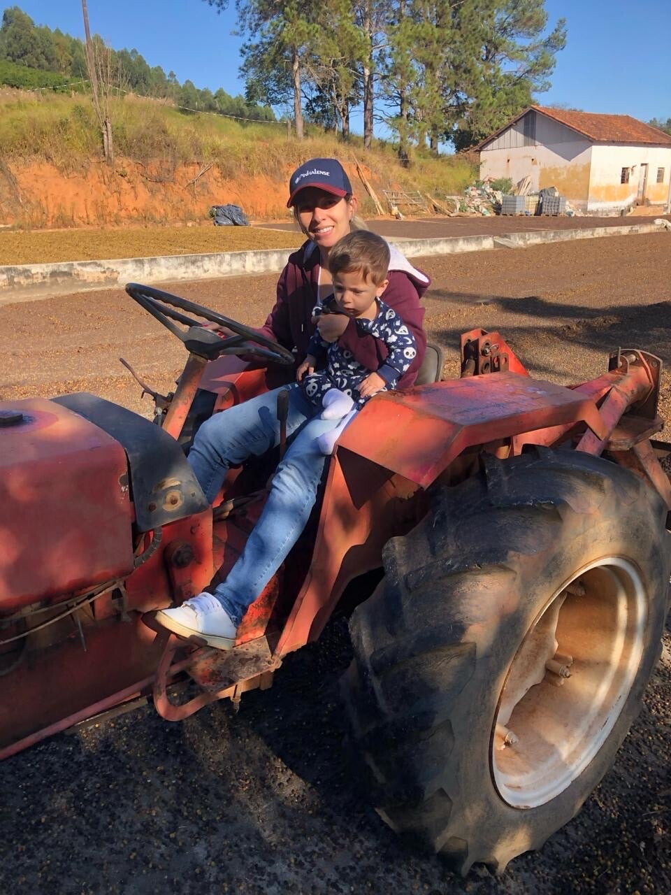 A woman and a young boy sitting on a red tractor in an outdoor farm setting with dirt tracks, trees, and a building in the background.