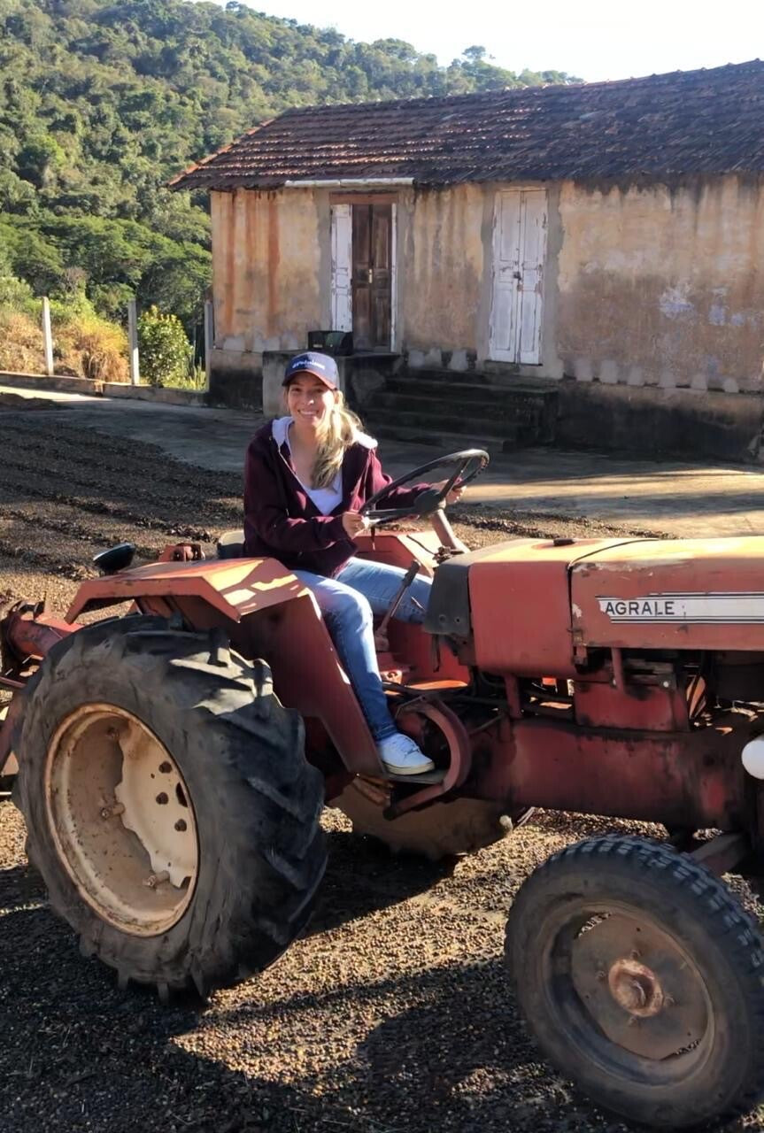 A woman sits on a red agricultural tractor in front of a rustic old building with a tiled roof, with a green hill in the background.