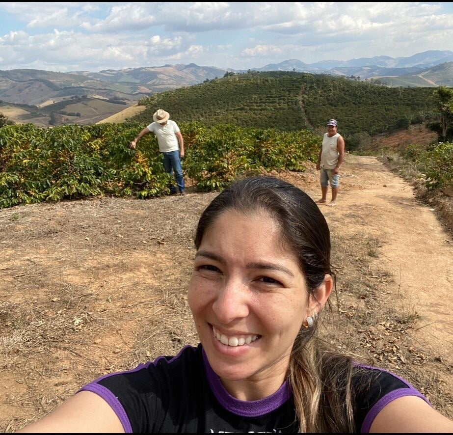 A smiling woman takes a selfie on a dirt path in a mountainous agricultural landscape, with two people working among crop plants in the background.